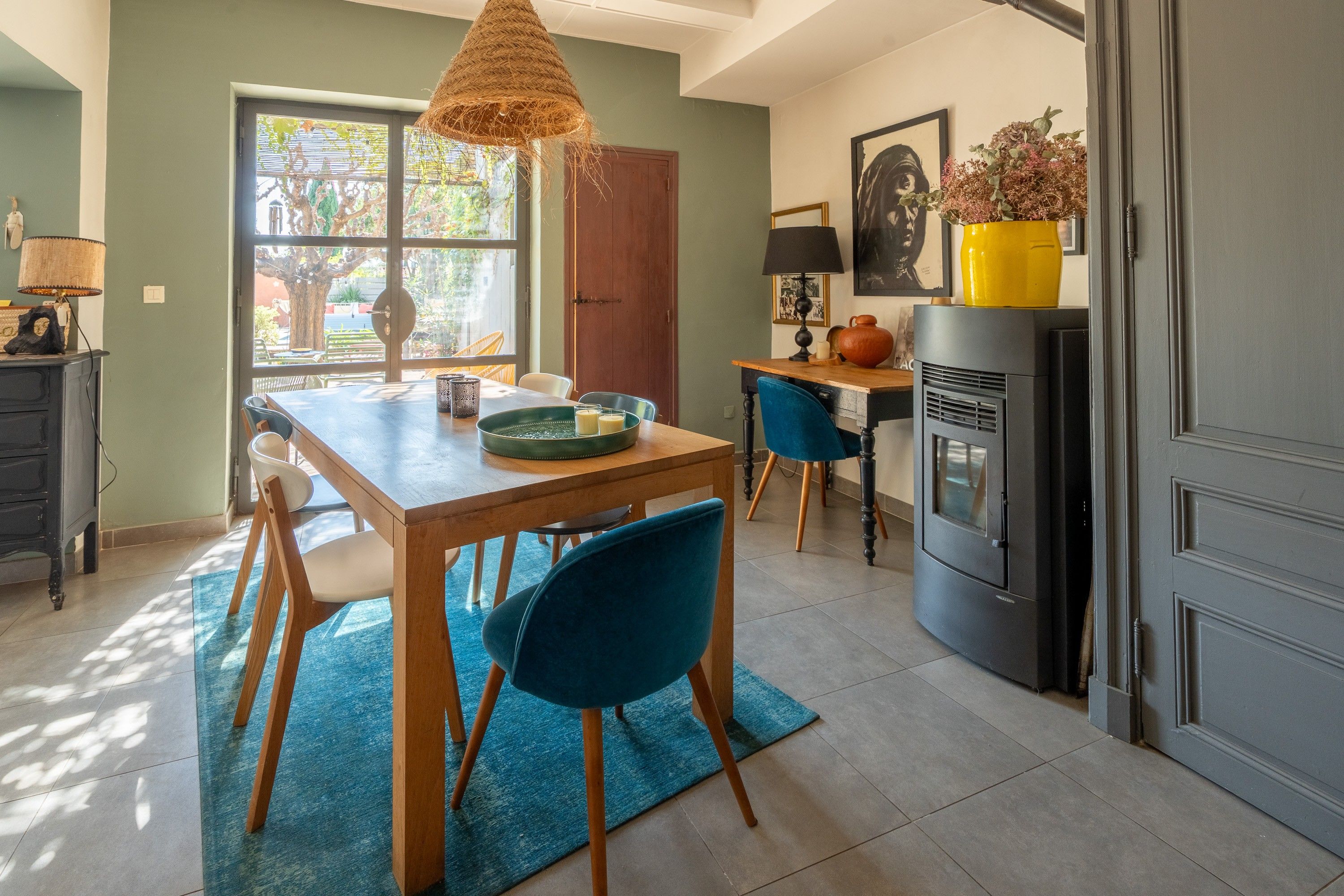 Un coin repas avec une table en bois, des chaises bleues, un luminaire suspendu en fibres tressées et une vue sur un espace extérieur, visible à travers une fenêtre.
