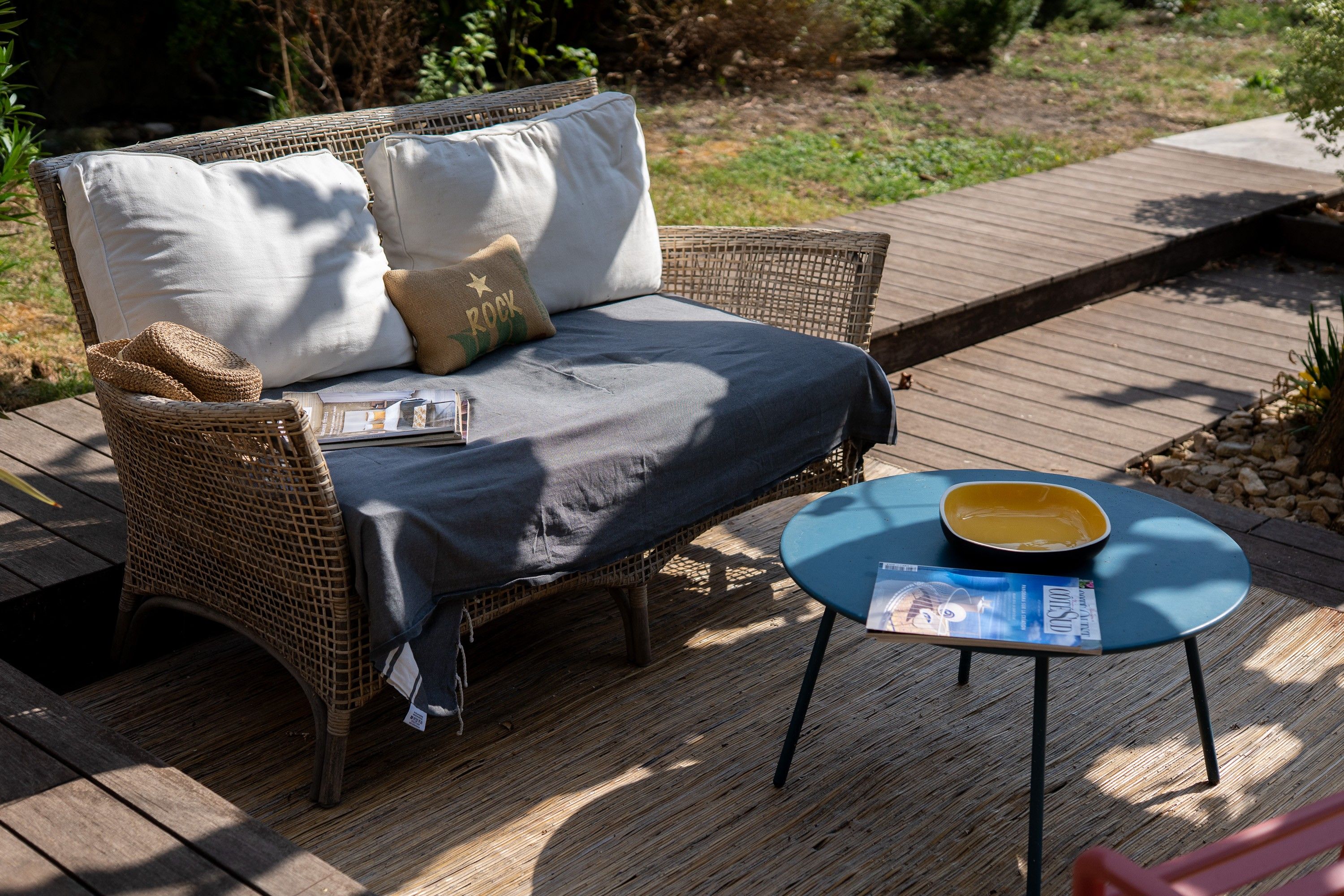 Un sofá de mimbre con cojines y una mesa azul con una revista encima, sobre una terraza de madera.