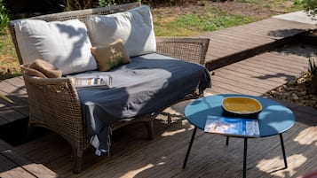 Un sofá de mimbre con cojines y una mesa azul con una revista encima, sobre una terraza de madera.