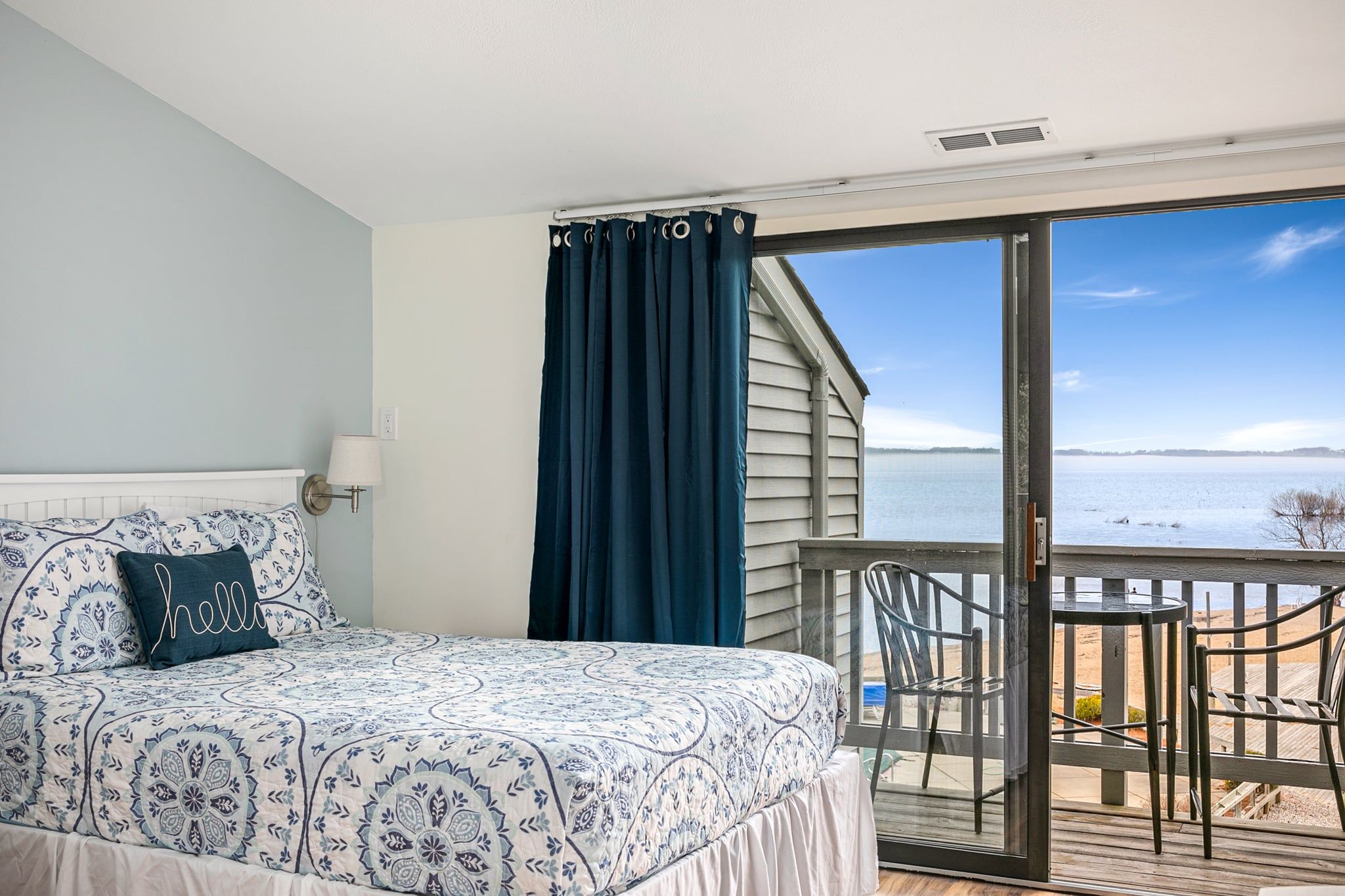 Un dormitorio con cama, mesita de noche, lámpara y vista a un cuerpo de agua a través de puertas corredizas de vidrio.