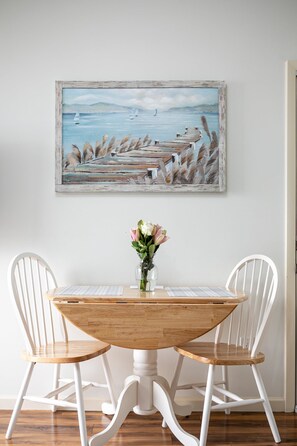 A dining area with a wooden table and white chairs, a painting above the table, and a vase with flowers on the table. - 🏖 Top-floor Bay-view 1BR @ The Shores Grand Traverse Resort Sleeps 6 (Williamsburg)