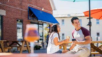 Two people sitting at an outdoor table with drinks, a brick building, and a blue awning in the background.