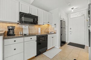 Una cocina con muebles blancos, un microondas negro, una cafetera, un lavavajillas y un refrigerador.