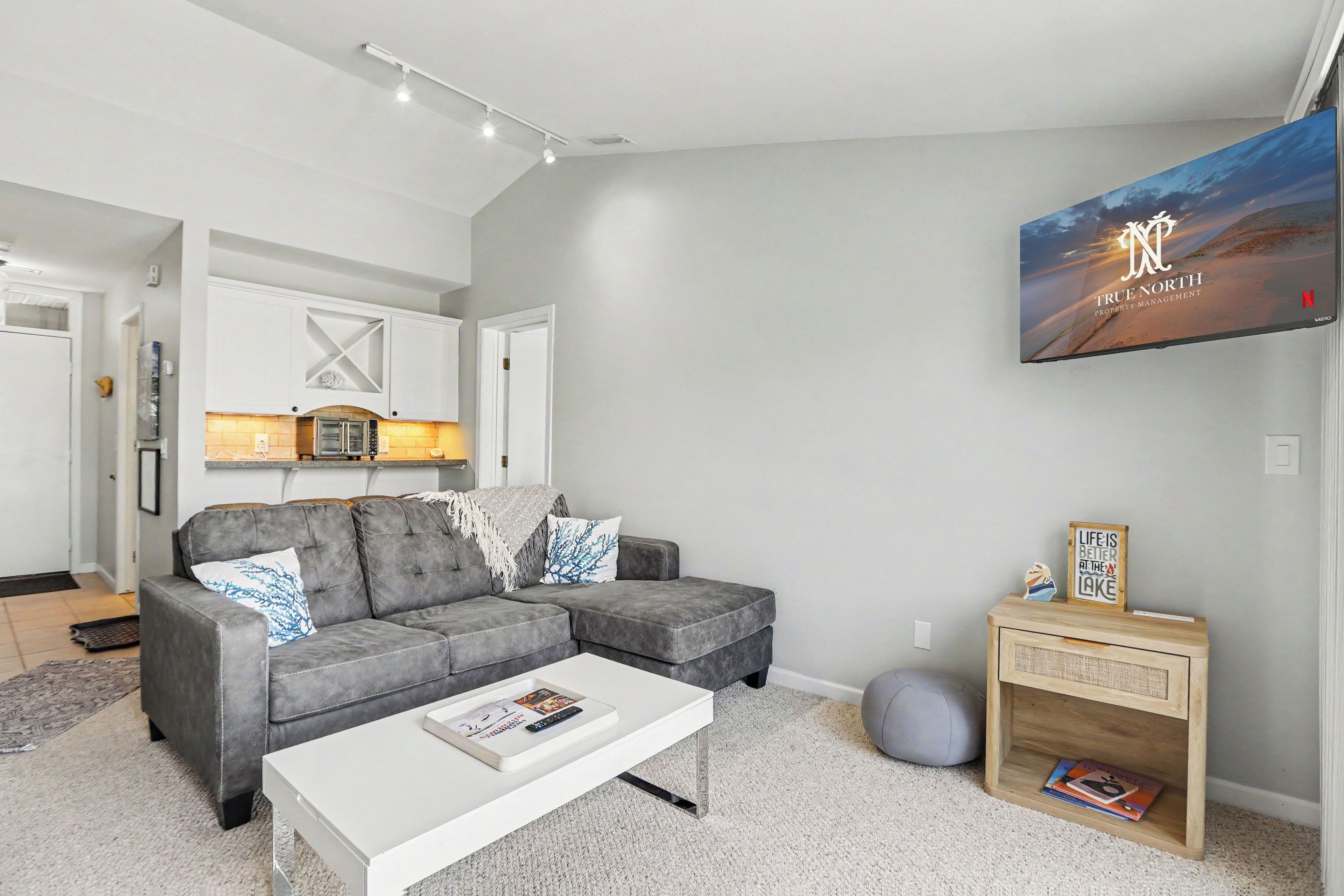 A modern living room with a grey sectional sofa, a white coffee table, and a mounted TV displaying 'True North Property Management'.