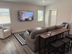 A modern living room with a large sofa, a coffee table, and a TV displaying a beach scene. - New Construction.  Sunny Escape Near Anna Maria : 3BR/2BA Home with Resort Pool! (Bradenton)