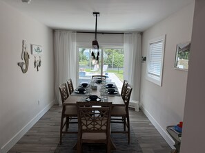 Un coin repas avec une table dressée pour six personnes, des chaises, un luminaire suspendu et une vue sur un jardin par la fenêtre.
