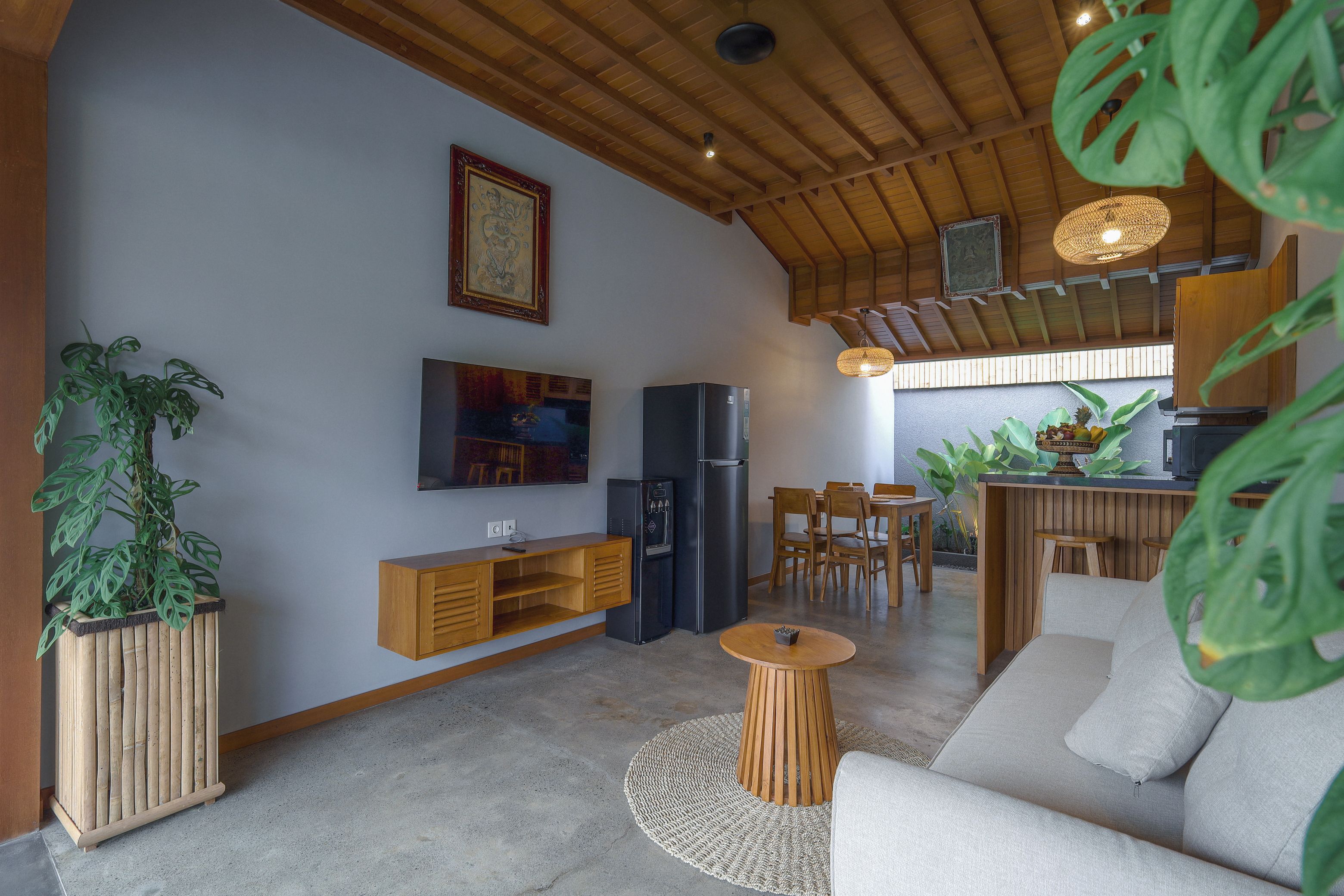 Un espacio habitable moderno con techo de madera, televisor de pantalla plana, refrigerador, zona de comedor con sillas de madera y plantas en macetas.