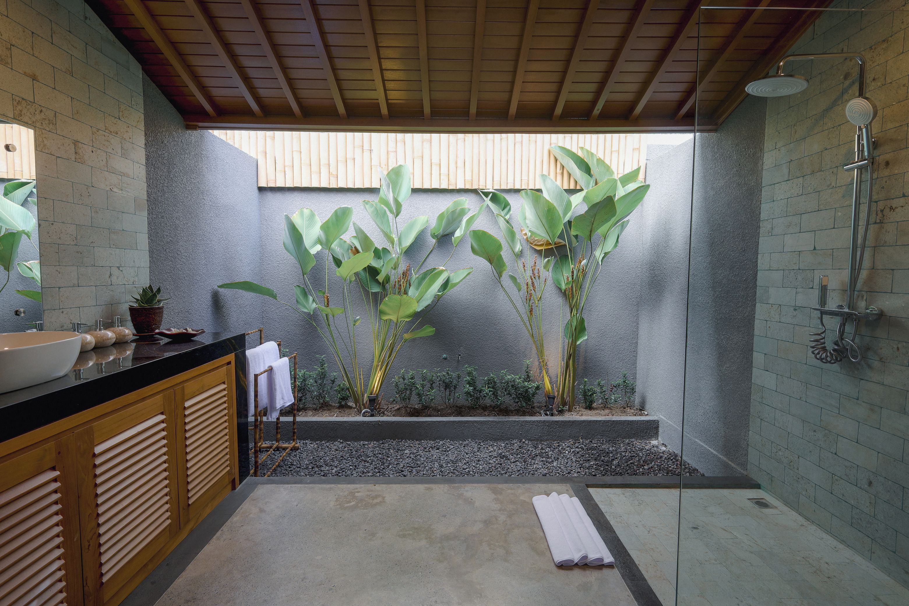 Un baño moderno con un lavamanos negro, armarios de madera, ducha y plantas verdes grandes.
