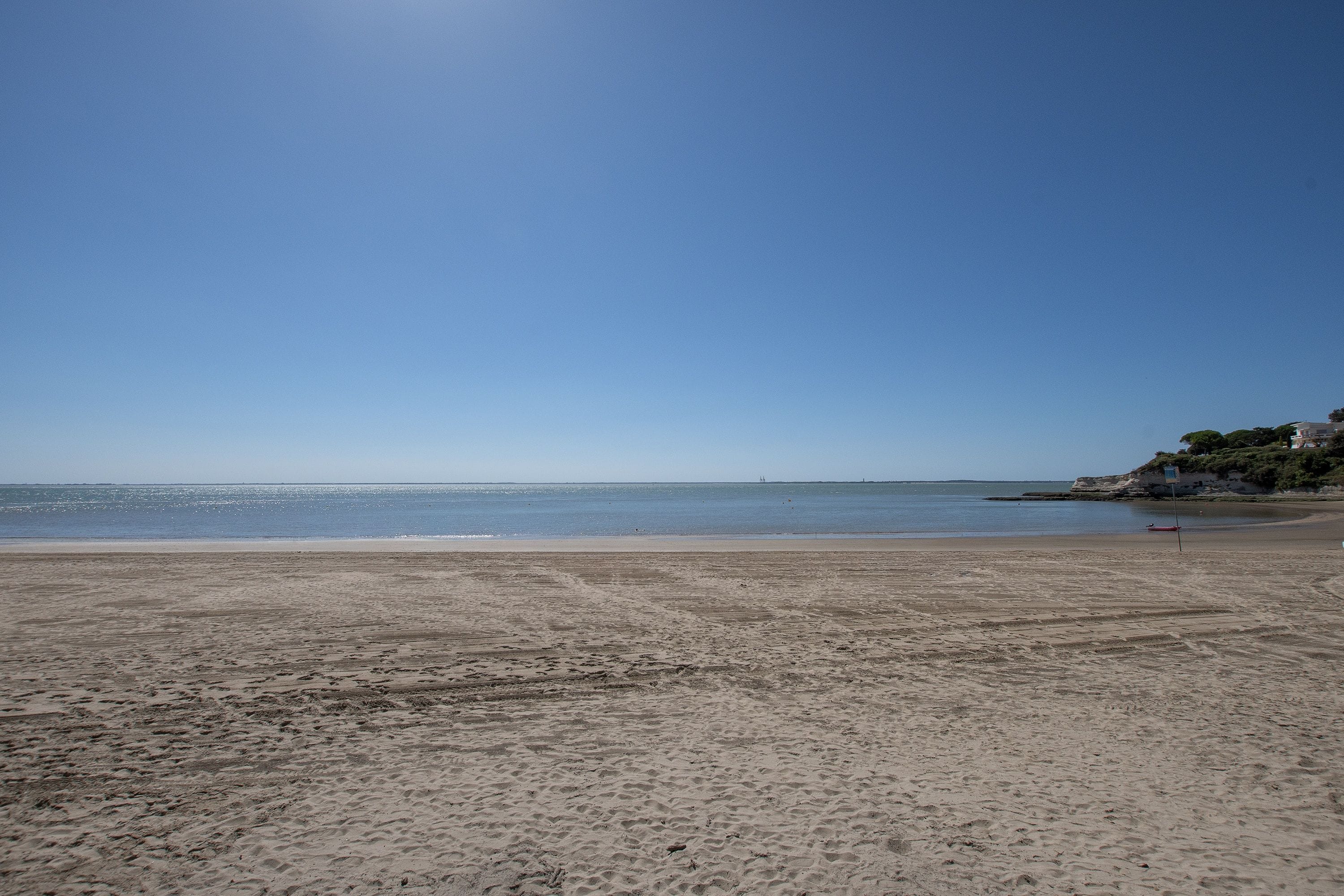 Ein Strand mit blauem Himmel, ruhiger See und einem Felsen rechts.
