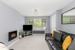 A modern living room with a fireplace, a flat-screen TV, a leather sofa, and a large window with blinds. - Maison Modern - Family-Friendly with Lovely Garden - Pass the Keys (Leeds)