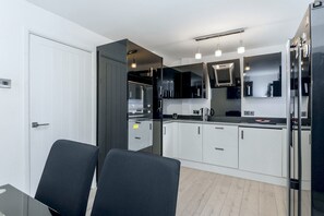 A modern kitchen with white cabinets, a black countertop, and a built-in oven. There are two chairs at a table, and a refrigerator is visible on the right. - Maison Modern - Family-Friendly with Lovely Garden - Pass the Keys (Leeds)