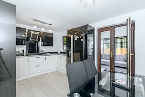 A modern kitchen with white cabinets, a black countertop, and stainless steel appliances. There is a dining area with a glass table and a gray chair. - Maison Modern - Family-Friendly with Lovely Garden - Pass the Keys (Leeds)