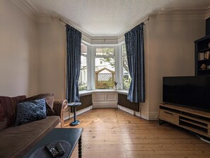 A living room with a wooden floor, a sofa, a coffee table, a TV stand, and a bay window with curtains. - The Seaside Family Retreat - Pass the Keys (Whitley Bay)