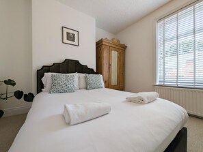 Een net opgemaakt bed met kussens en een opgevouwen handdoek, een houten garderobe en een raam met jaloezieën.