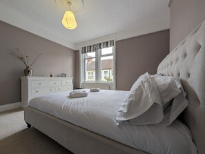 A bedroom with a large bed, a dresser, a window with curtains, and a hanging light fixture. - The Seaside Family Retreat - Pass the Keys (Whitley Bay)
