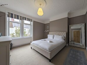 Een slaapkamer met een groot bed, een nachtkastje, een hanglamp, een raam met uitzicht en een spiegel met een versierde lijst.