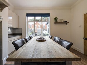 A dining area with a long wooden table, black leather chairs, a window with a view of a brick building, and a shelf with a plant and decorative items. - The Seaside Family Retreat - Pass the Keys (Whitley Bay)