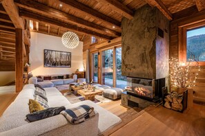 A cozy living room with a fireplace, wooden beams, a large window, and a sofa set. - Chalet Twin - Piscine Privée et Sauna (La Salle-les-Alpes)