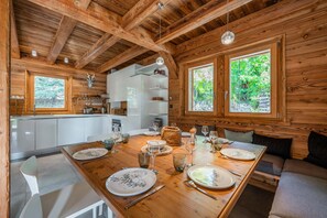 A wooden cabin interior with a dining table set for four, kitchen cabinets, and a large window overlooking greenery.