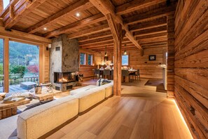 A log cabin interior with a stone fireplace, wooden dining area, and large windows offering a view of the outdoors.