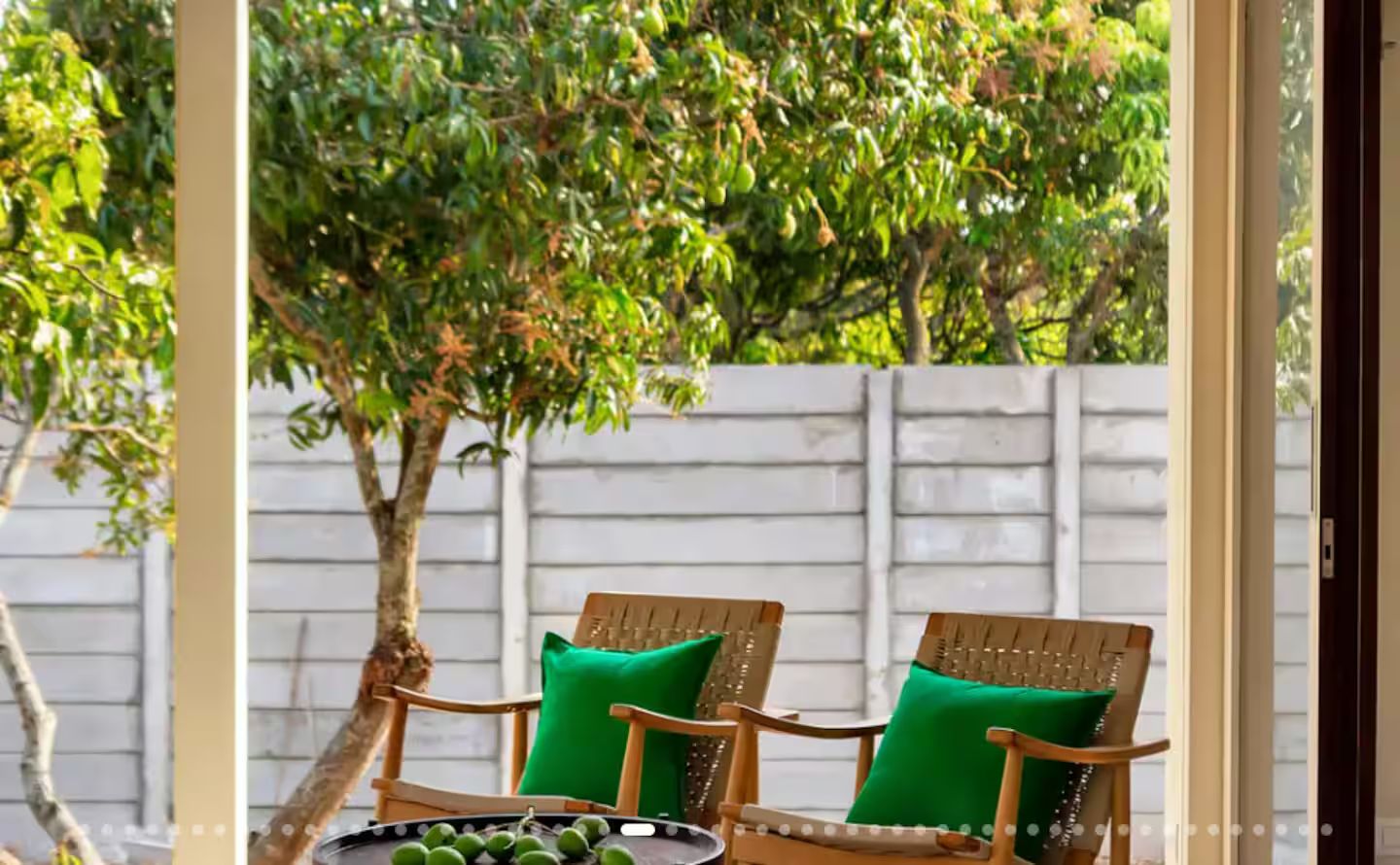 Two wooden chairs with green cushions, a metal tray with fruits, and a white fence in the background.