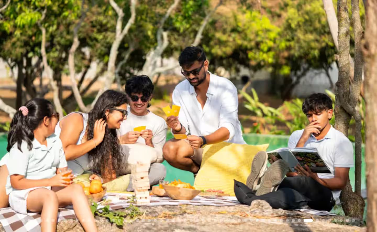 A group of people sitting on a checkered blanket outdoors, some eating and others reading.