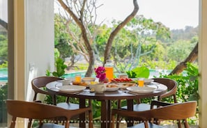 Una mesa de comedor con platos, cubiertos y un vaso de jugo de naranja, frente a un fondo de vegetación exuberante y una piscina.