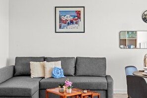 Un canapé gris avec des coussins, une petite table d’appoint en bois sur laquelle est posée une plante, et un tableau encadré accroché au mur.