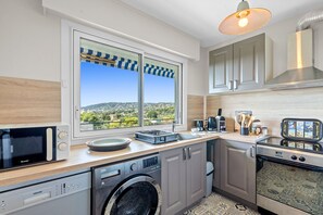 A modern kitchen with a large window, built-in appliances, and a view of the landscape. - Le Panoramic Lérins - 2km de la Plage (Antibes)