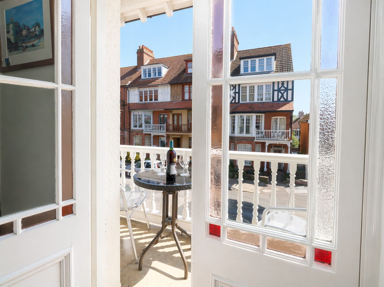 A room with a view of a street, a table with a bottle and glasses, and a window with a white frame.