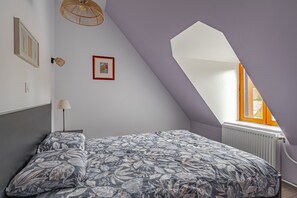 A bedroom with a sloped ceiling, a bed with floral bedding, a framed picture, a floor lamp, and a window with a view.