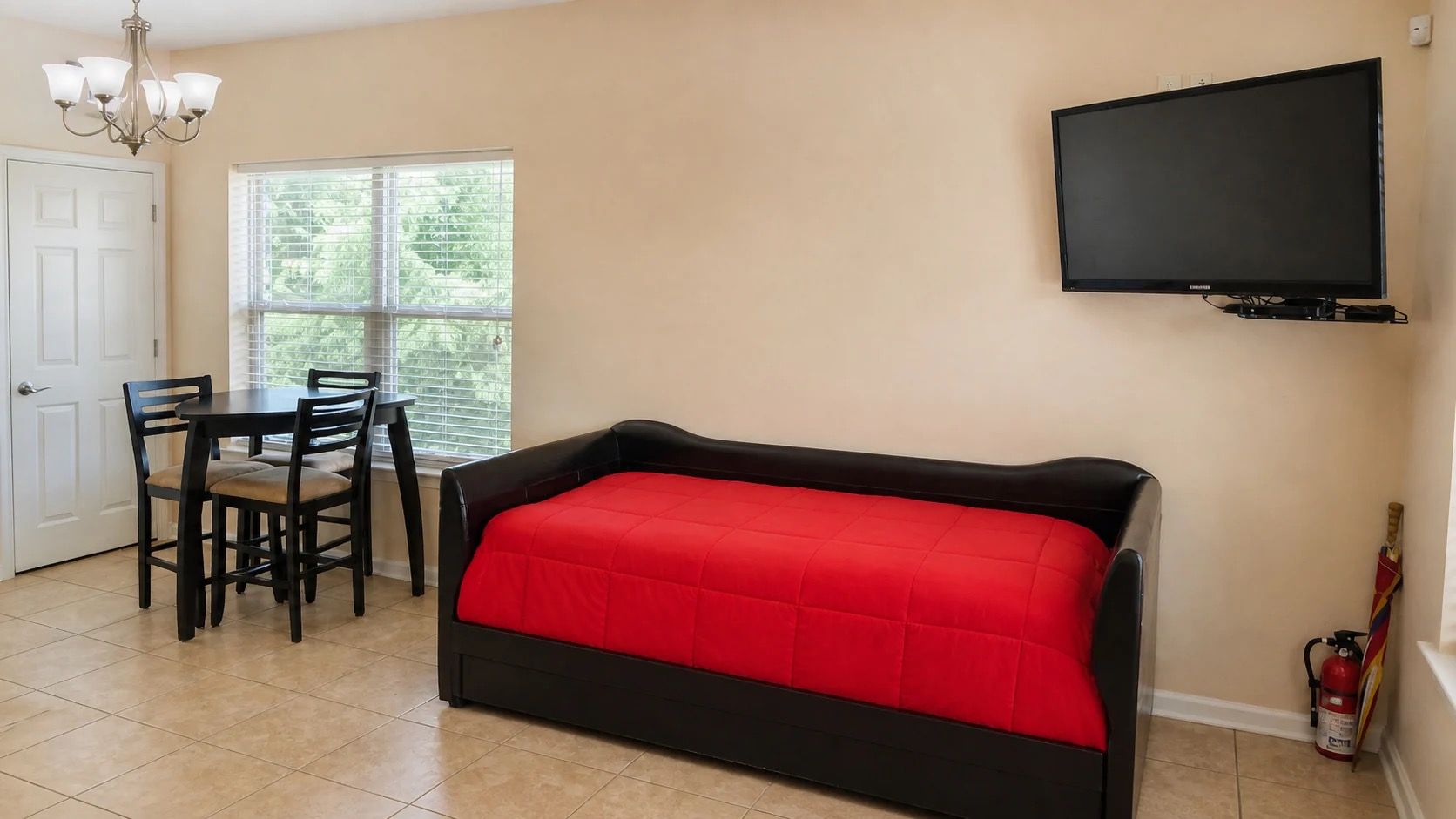 Une pièce comprenant un lit, un téléviseur fixé au mur, une table à manger avec des chaises et une fenêtre avec des stores.