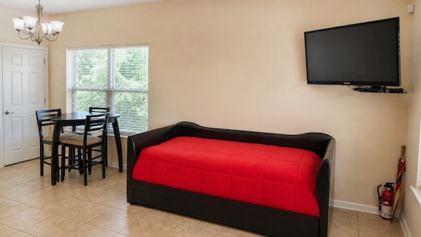 A room with a bed, a TV mounted on the wall, a dining table with chairs, and a window with blinds.