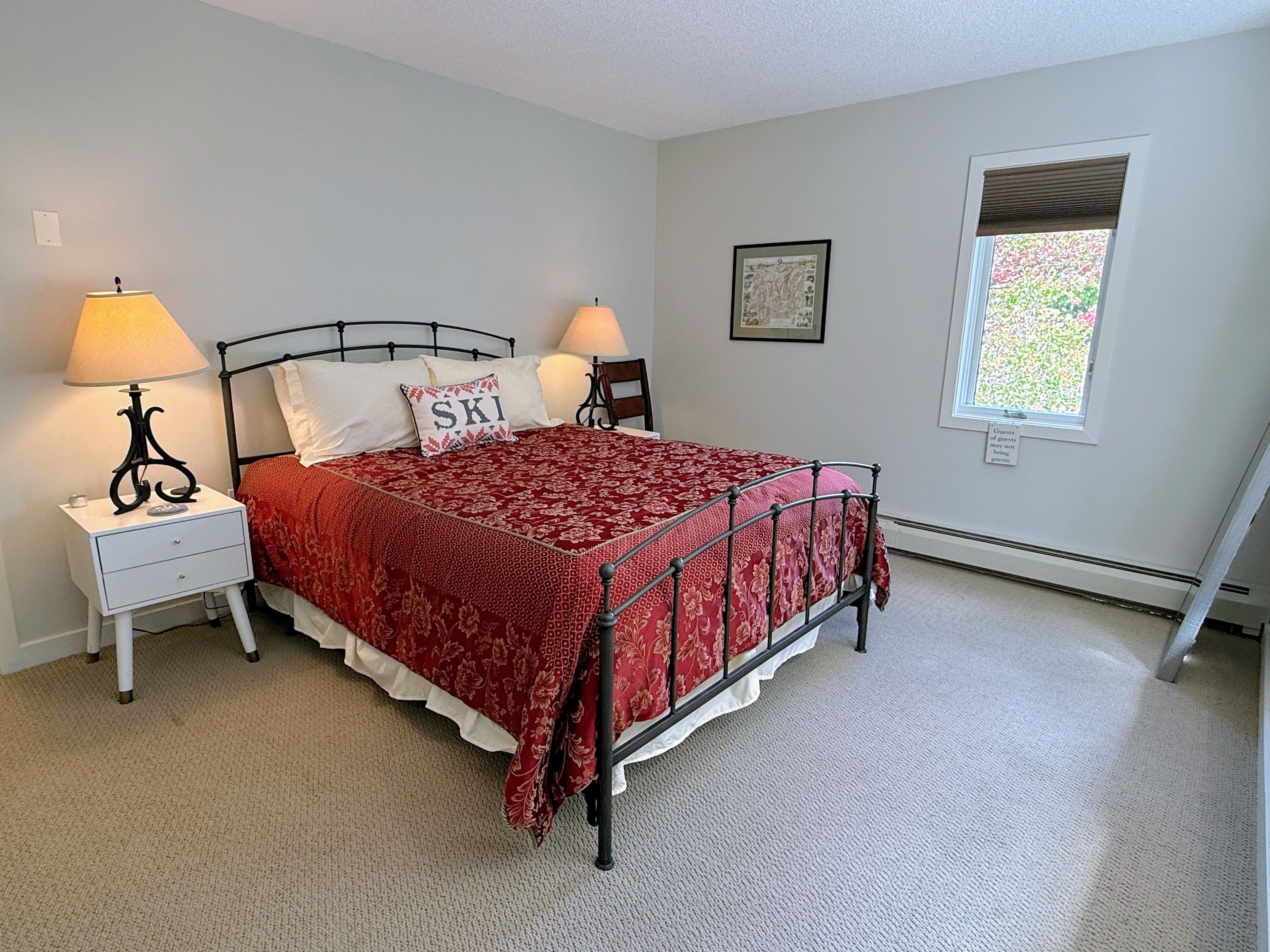 A bedroom with a bed, two bedside tables, lamps, a window, and a framed picture on the wall.
