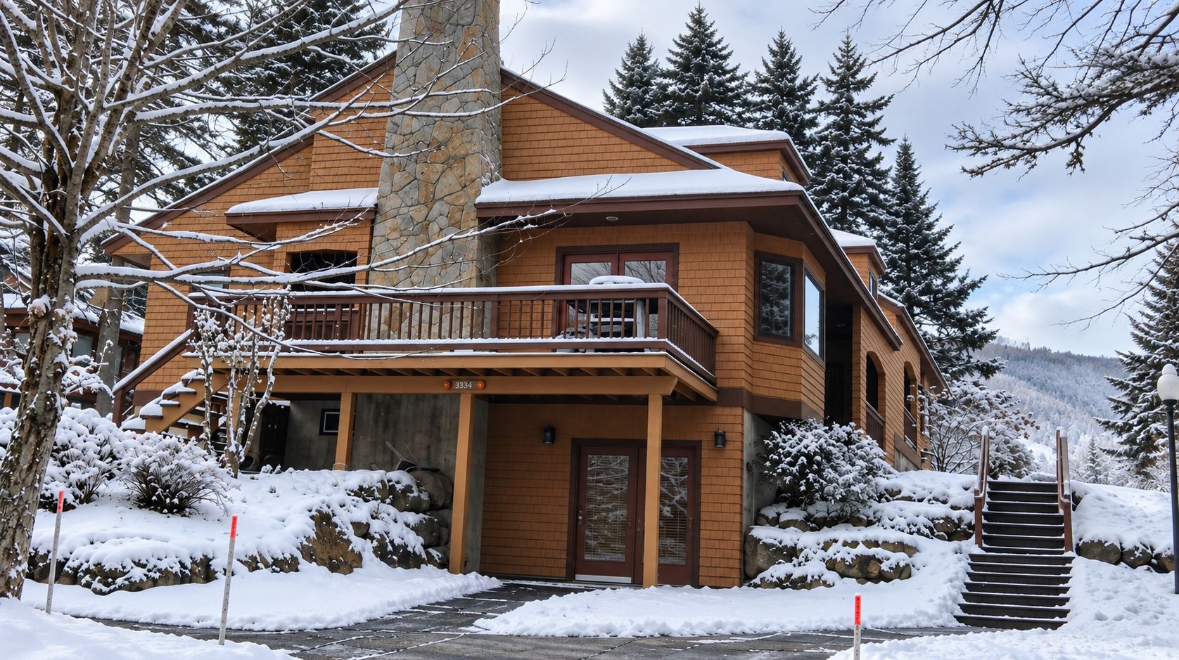 Una casa de madera de dos pisos con un balcón, rodeada de árboles cubiertos de nieve y una montaña al fondo.