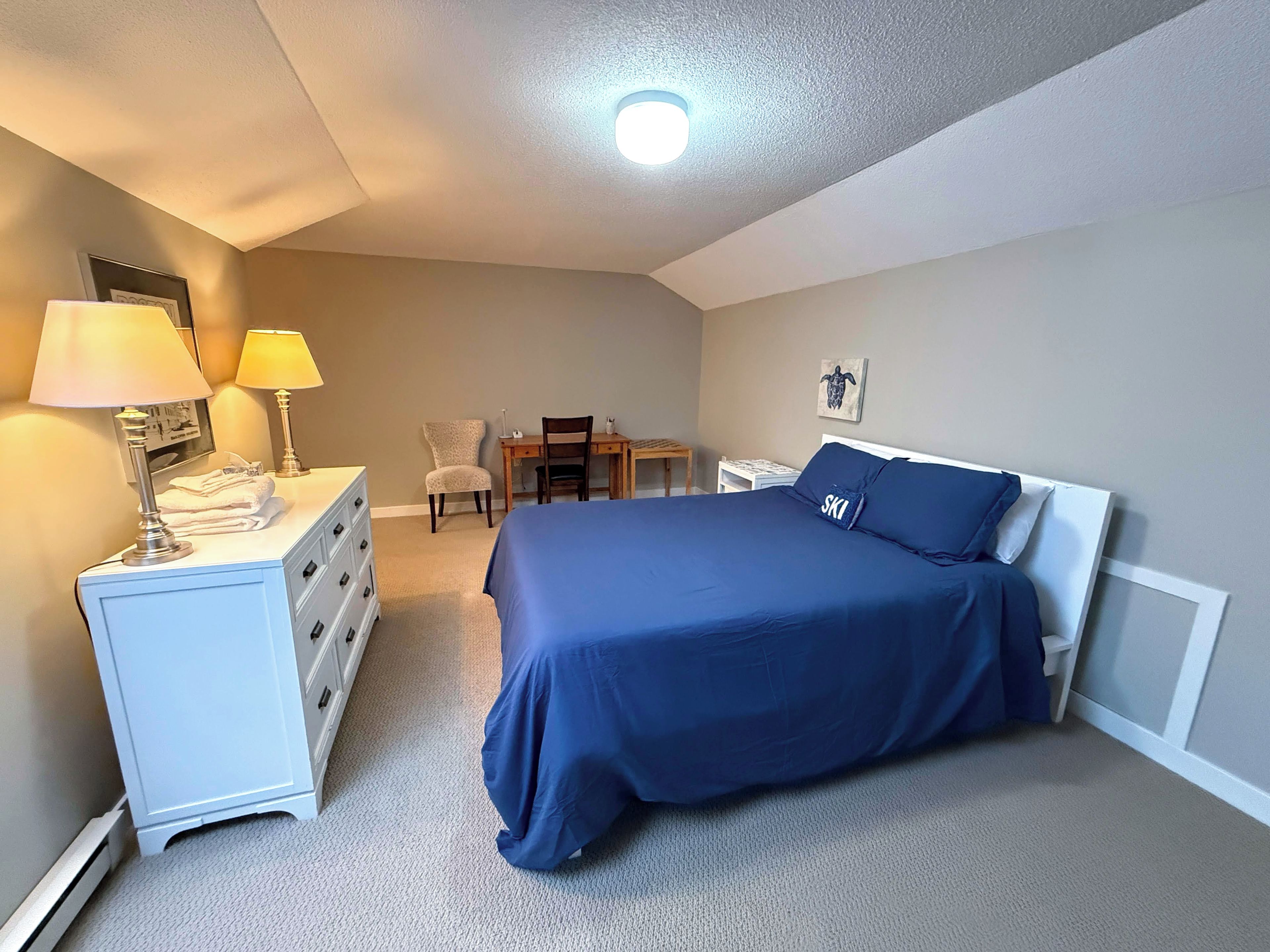 A bedroom with a bed, a desk, two chairs, a dresser, and a lamp.