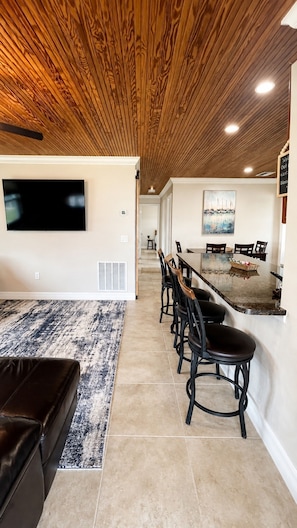 A modern living room with a flat-screen TV, a dining area with a long table and chairs, and a kitchen area with a marble countertop.