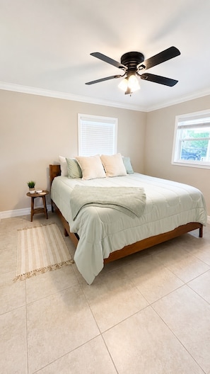 A bedroom with a bed, a ceiling fan, a small side table with a plant, and a window.