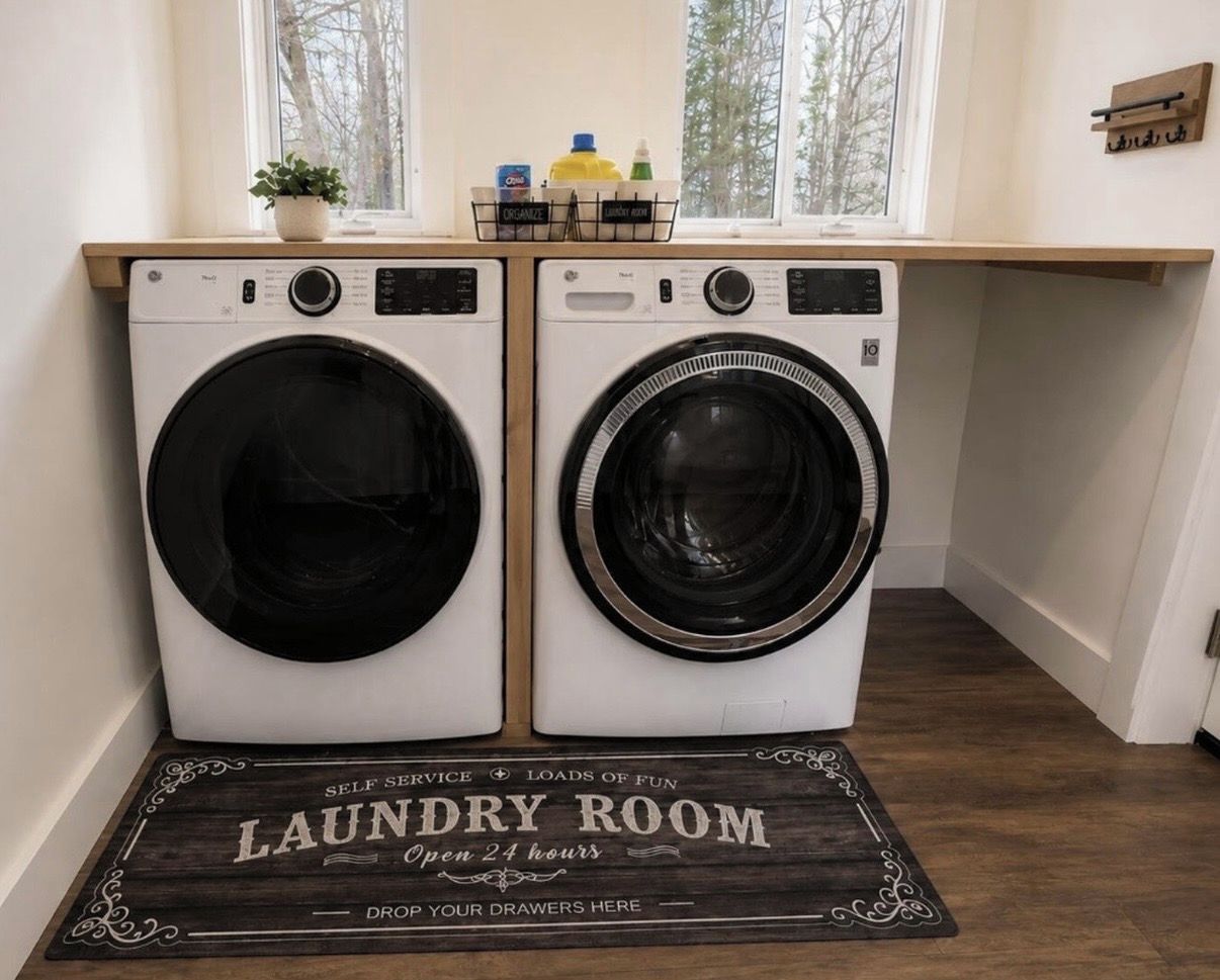 A laundry room with two washing machines, a wooden shelf with laundry supplies, and a doormat stating 'Laundry Room Open 24 hours.'