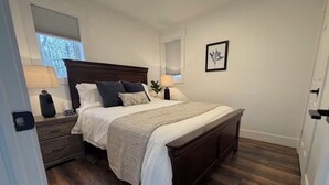 A bedroom with a wooden bed, two bedside tables, a nightstand, a window with blinds, and a framed picture on the wall.