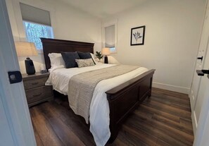 A bedroom with a wooden bed, two bedside tables, a nightstand, a window with blinds, and a framed picture on the wall.