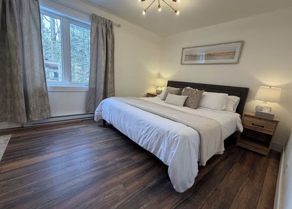 A bedroom with a large bed, wooden flooring, a window with curtains, and a wall-mounted light fixture.