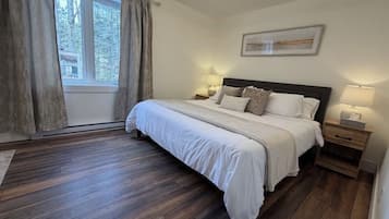 A bedroom with a large bed, wooden flooring, a window with curtains, and a wall-mounted light fixture.