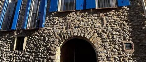 Un bâtiment en pierre doté d’une porte d’entrée en arc de cercle au centre et de nombreuses fenêtres avec des volets bleus.
