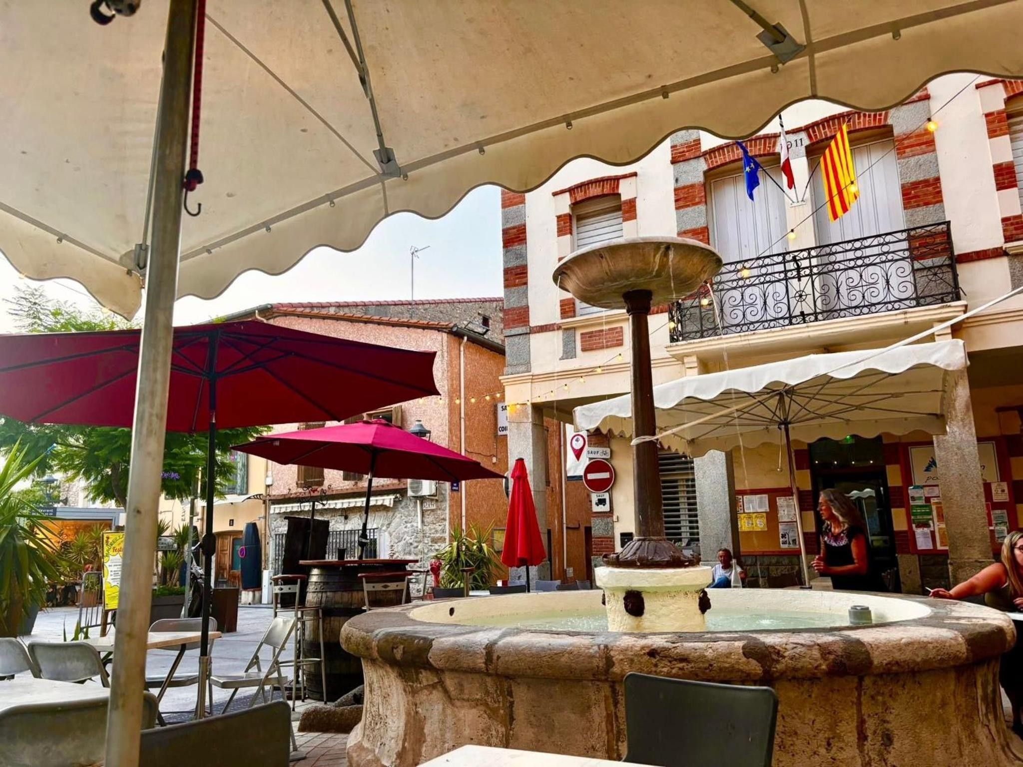 An outdoor café with tables and chairs, a fountain, and a building with balconies and flags.