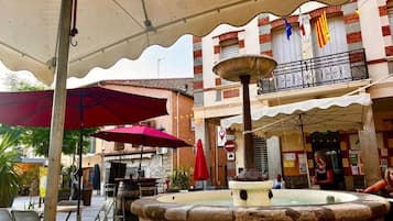An outdoor café with tables and chairs, a fountain, and a building with balconies and flags.