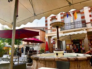Un café al aire libre con mesas y sillas, una fuente y un edificio con balcones y banderas.