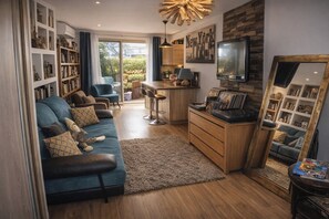 A living room with a blue sofa, a coffee table, a TV, a bookshelf, and a dining area with a bar stool. - Cozy 2-room apartment on the first floor in a quiet complex with a pool (Antibes)