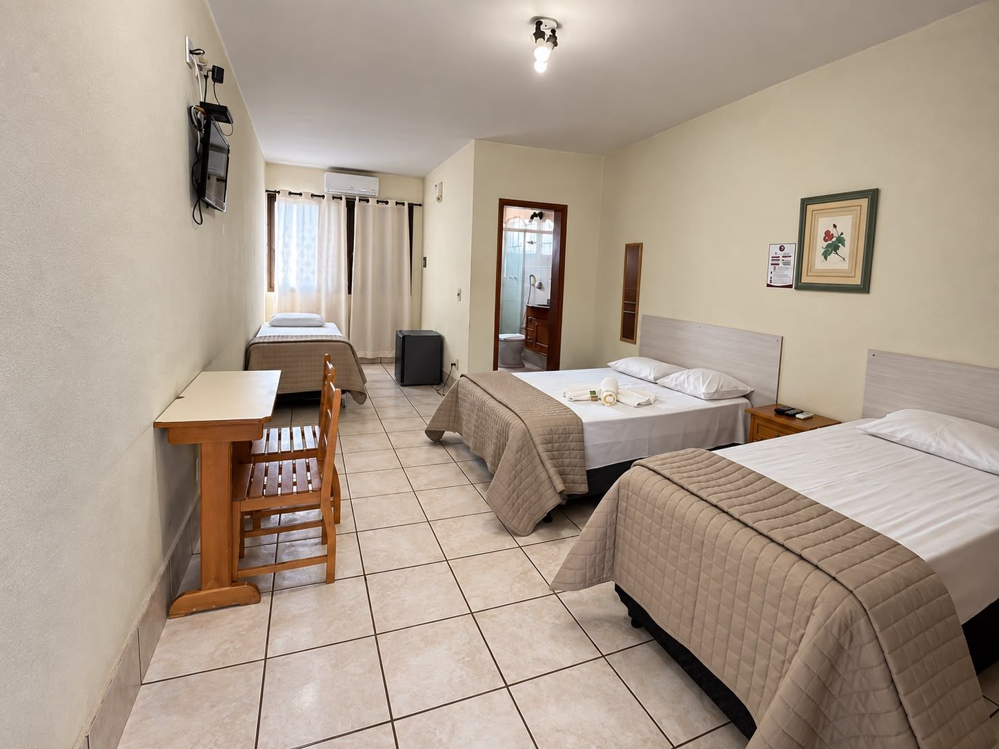 A hotel room with two beds, a desk, a chair, a small table, a mirror, a picture frame, and a wall-mounted light fixture.