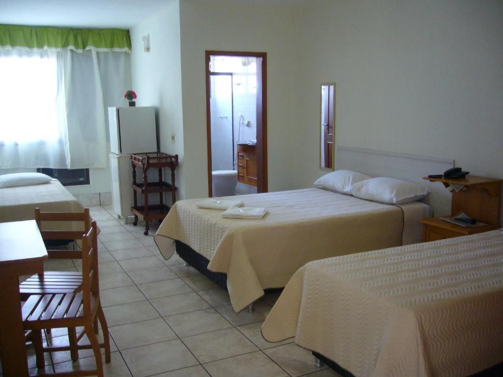Quarto de hotel com duas camas, uma mesa pequena, uma cadeira, um refrigerador e banheiro visível por uma porta aberta.